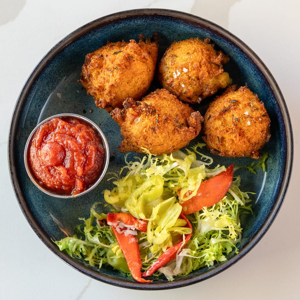 Four crispy golden fritters served with tomato dipping sauce, mixed greens, sliced peppers, and tomatoes on a dark ceramic plate.