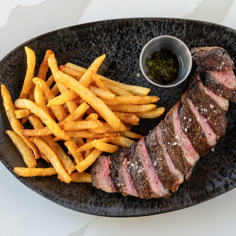 Sliced grilled steak with golden fries and green dipping sauce on a black oval plate.
