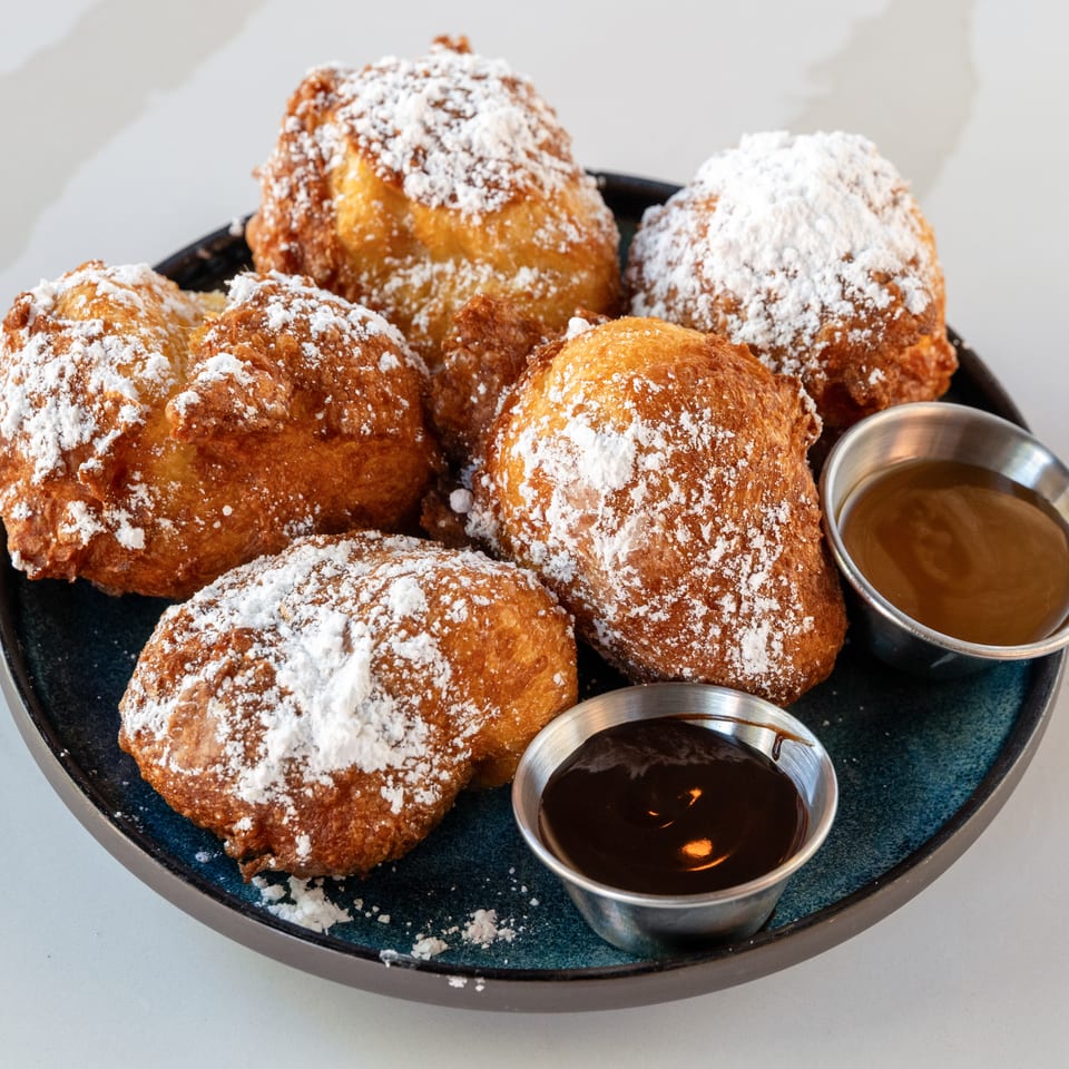 Golden fried pastries dusted with powdered sugar on a black platter, served with cups of caramel and chocolate dipping sauces.