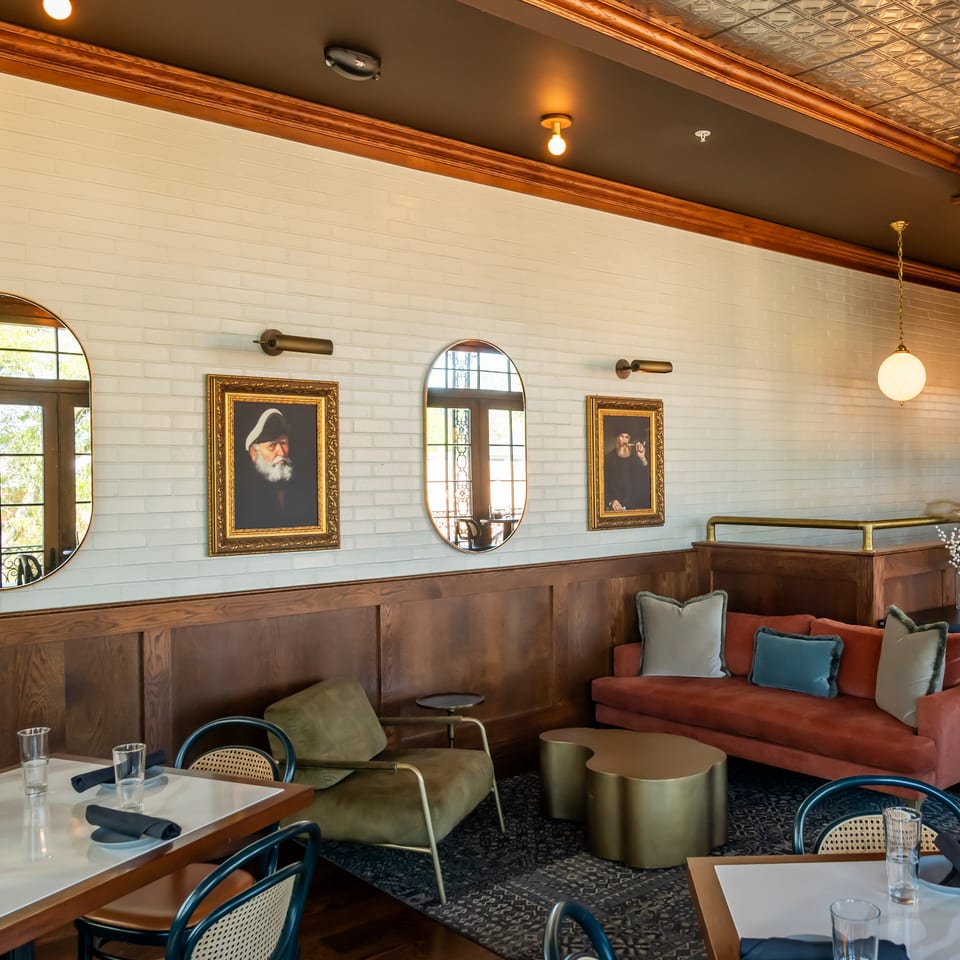 Elegant restaurant dining room with white brick walls, portrait artwork, arched mirrors, wood paneling, and colorful lounge seating.