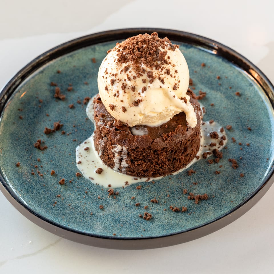 Chocolate brownie topped with melting vanilla ice cream and chocolate crumble on a blue ceramic plate.