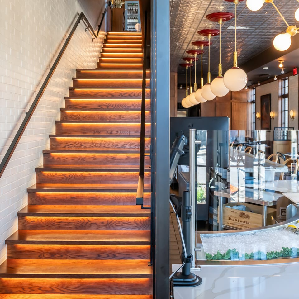 Upscale restaurant interior with illuminated wooden staircase, globe pendant lights, dining area, and glass seafood display counter.
