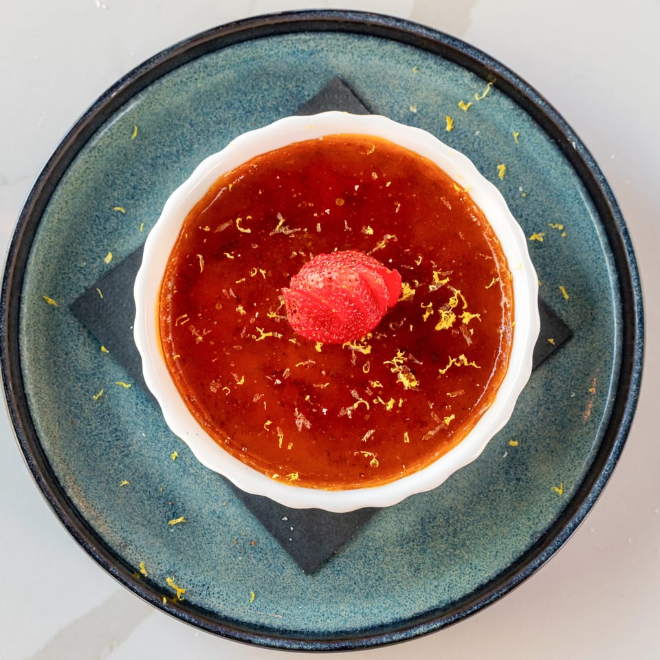 Caramelized dessert topped with sliced strawberries and lemon zest in a white dish on a blue ceramic plate.