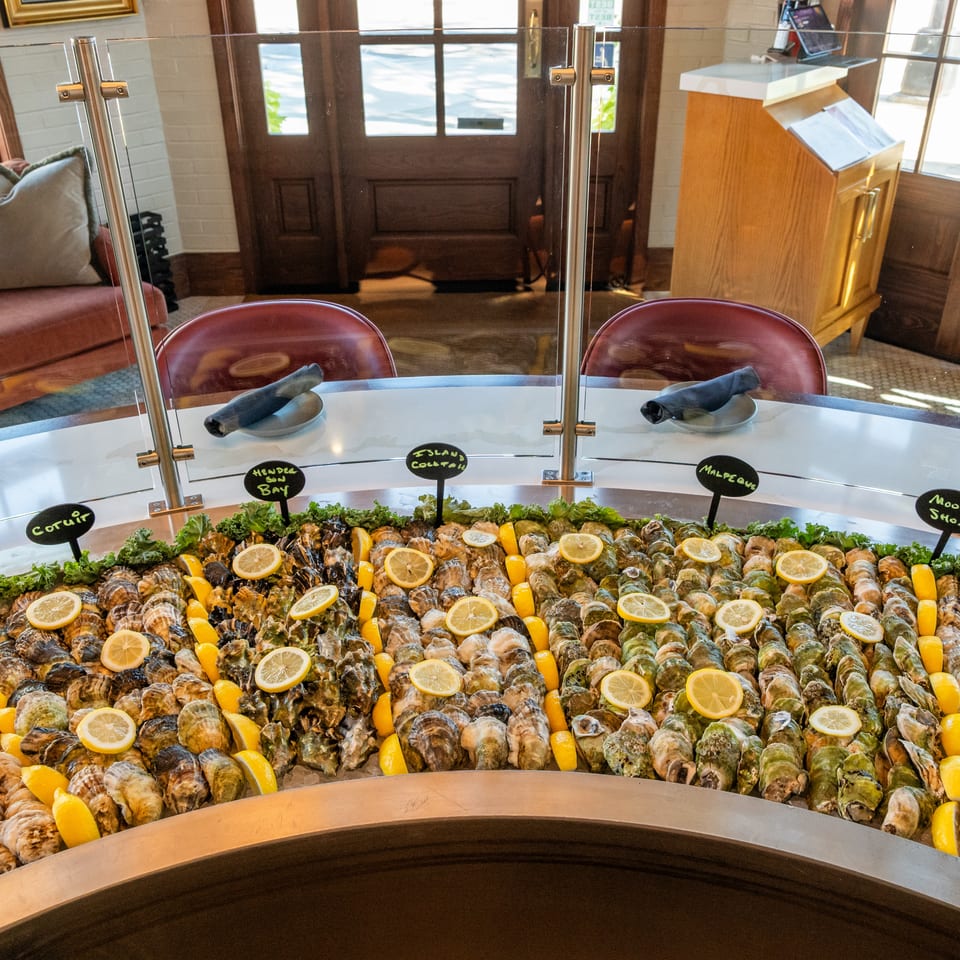 A restaurant oyster bar displays assorted oysters on ice with lemon wedges, labeled by location, behind a glass sneeze guard.