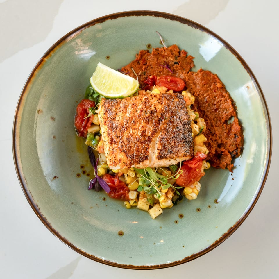 Seared fish fillet served with colorful vegetable relish, crispy fritters, microgreens, and a lime wedge on a ceramic plate.
