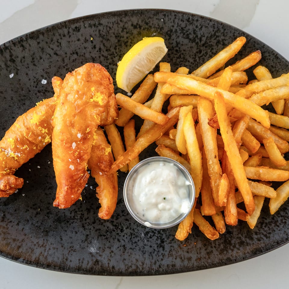 Crispy battered fish with golden fries, lemon wedge, and tartar sauce on a black plate.