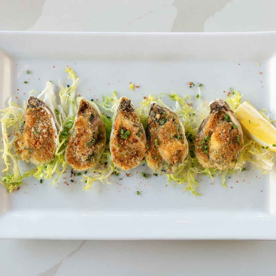 Five baked oysters topped with breadcrumbs and scallions, arranged on shredded lettuce with lemon wedges.