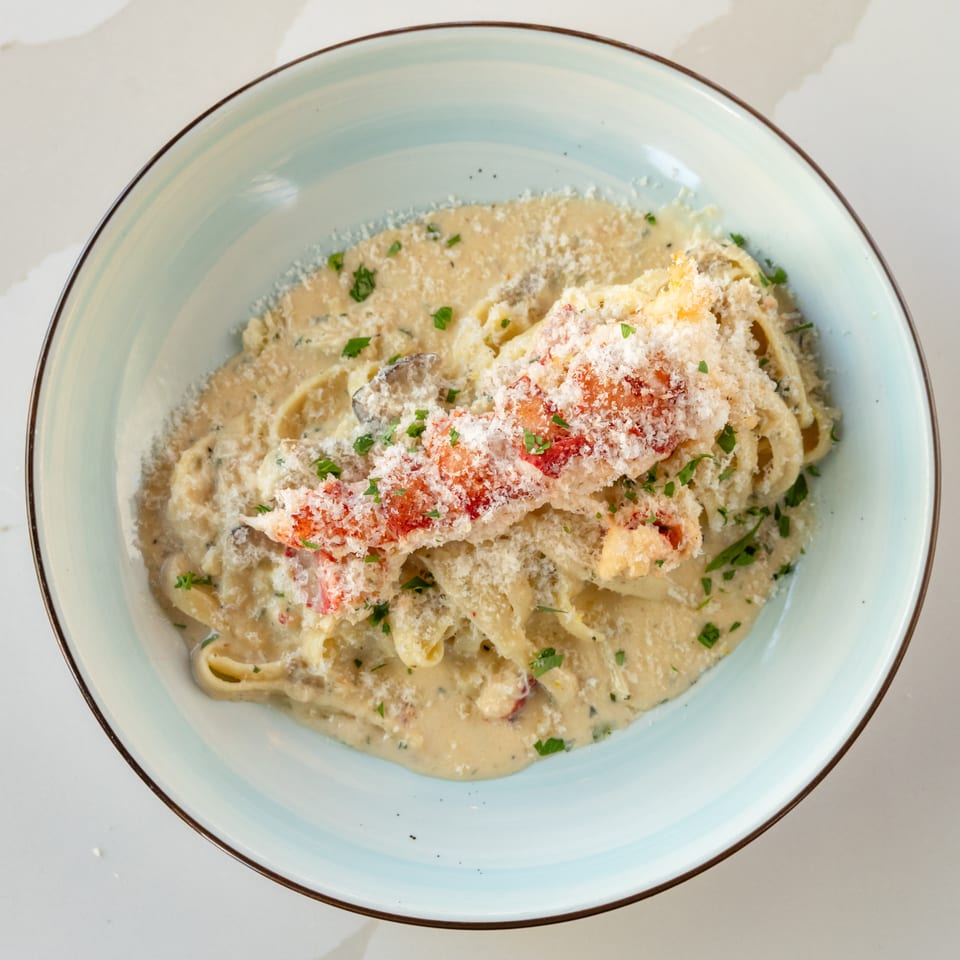 Creamy fettuccine pasta topped with lobster, grated Parmesan, red pepper, and parsley in a pale blue bowl.