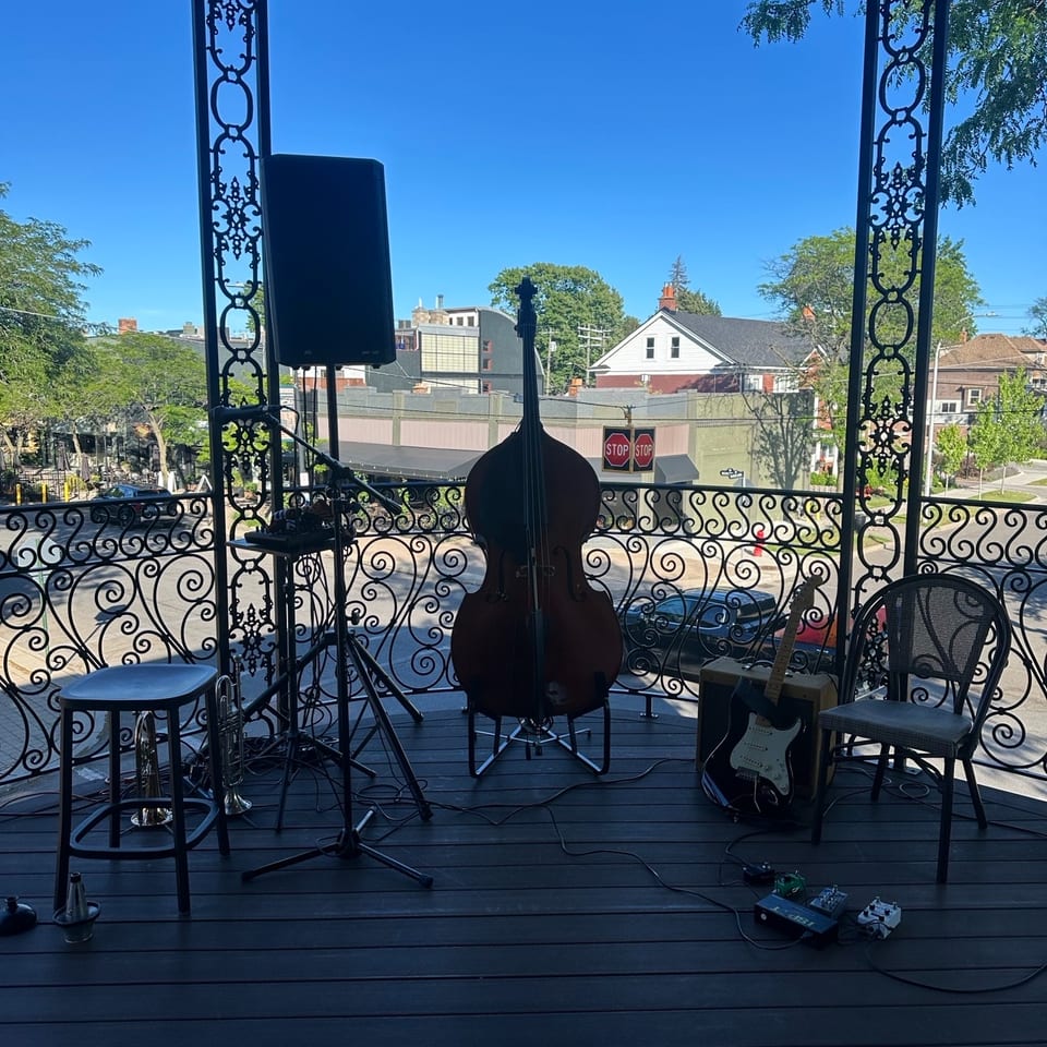 Outdoor gazebo stage with ornate ironwork, double bass, electric guitar, speakers, and microphone equipment overlooking a sunny neighborhood.