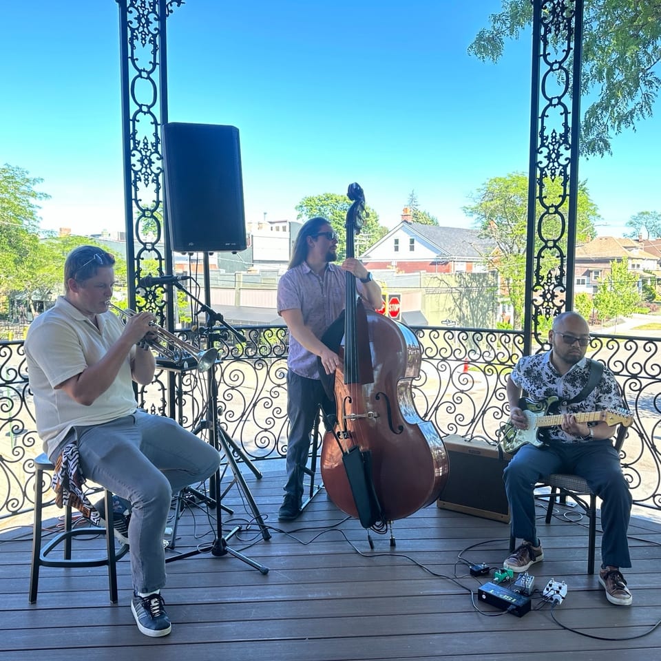 Three musicians perform on an ornate outdoor balcony, playing trumpet, double bass, and electric guitar against a sunny neighborhood backdrop.
