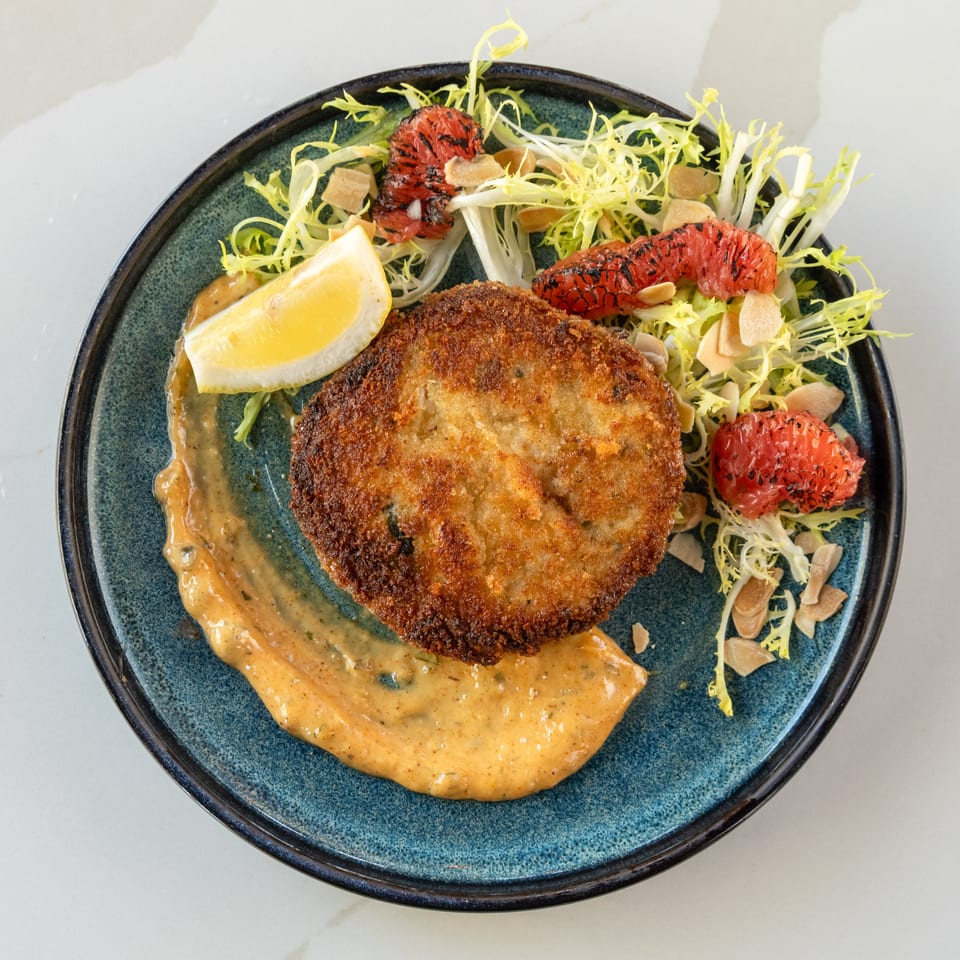 Crispy breaded cutlet served with lemon, creamy sauce, frisée salad, grilled grapefruit, and toasted almonds on a blue plate.