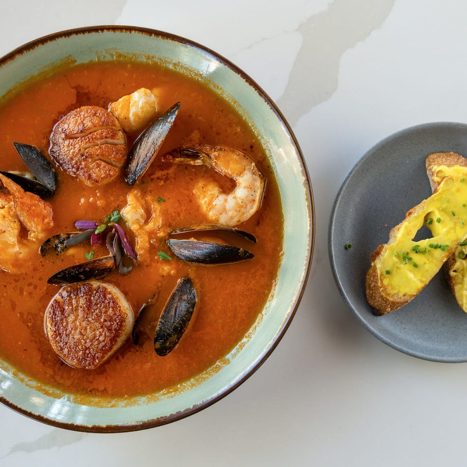 Overhead view of seafood stew with scallops, shrimp, and mussels beside cheesy toast on gray plates.