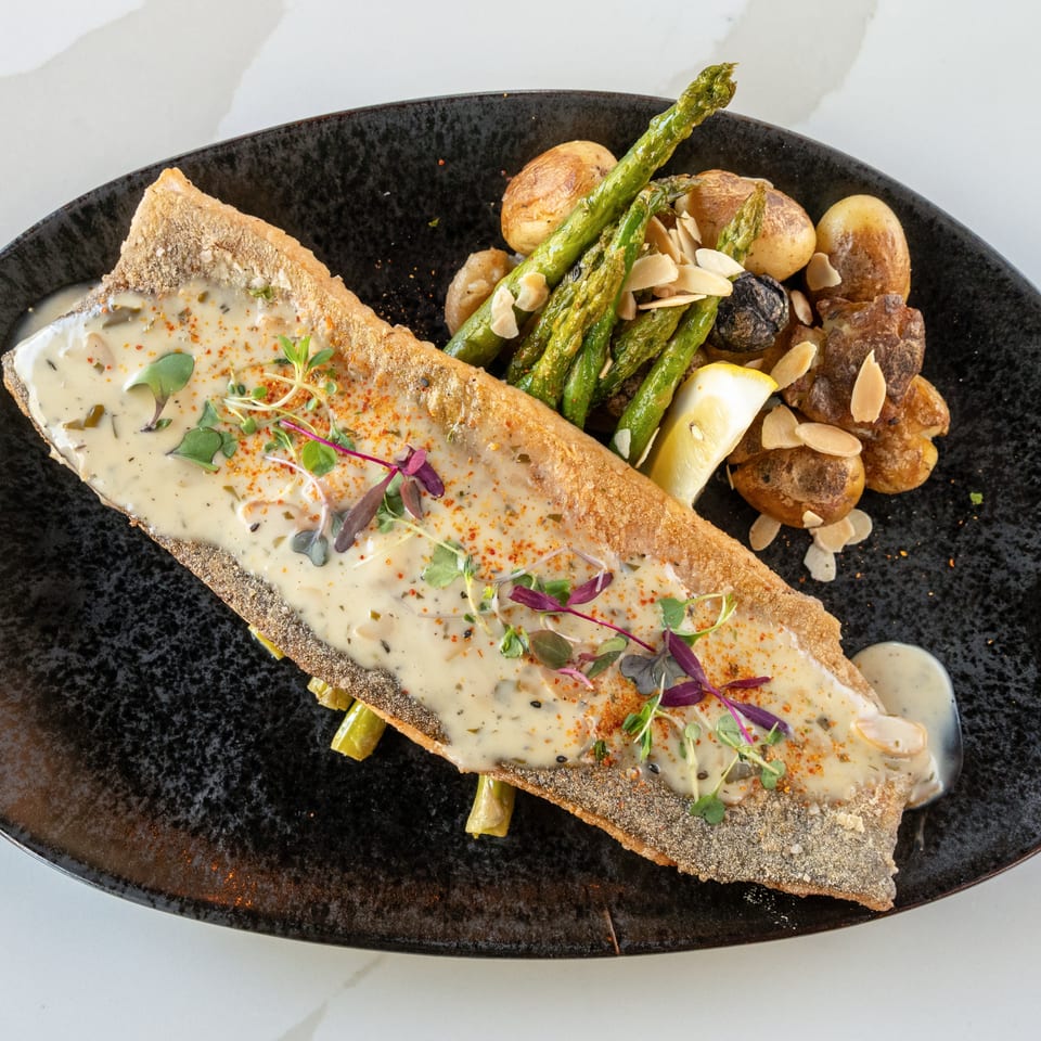 Crispy fish fillet with creamy herb sauce, roasted potatoes, asparagus, lemon, and almond slices on a black plate.