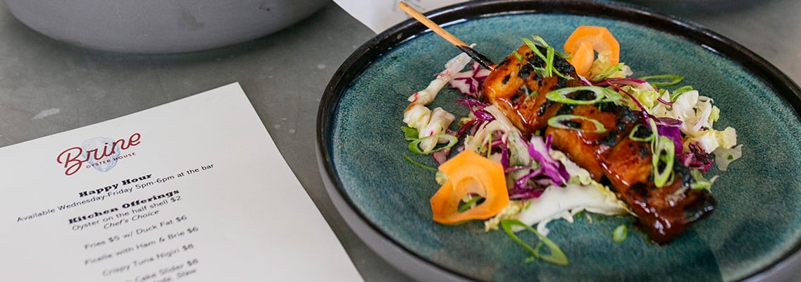 Glazed skewer dish on a teal plate beside the happy hour menu featured on the Brine Oyster House blog