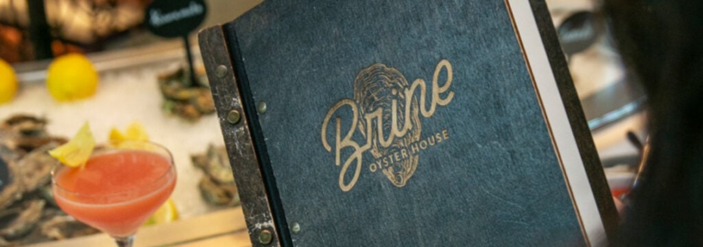 Brine Oyster House menu cover beside a pink cocktail and raw bar display on the contact Brine Oyster House page
